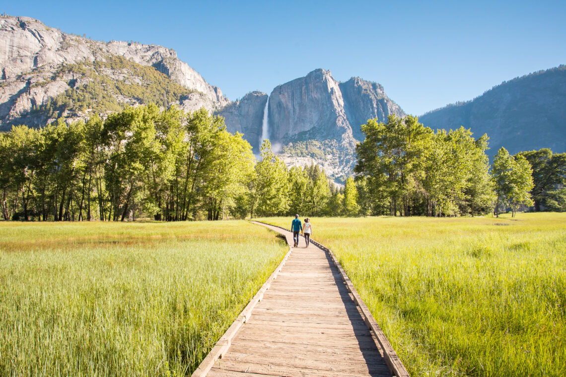 Wooden path through green grass in Sentinel Meadow with Yosemite Falls in the distance.